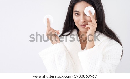 Skincare concept. Portrait of young happy asian woman in white bathrobe holding cotton pad covering her eye with it while standing on white background, smiling at camera and enjoying beauty routine Royalty-Free Stock Photo #1980869969