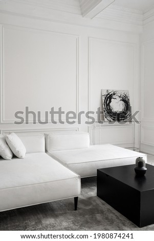Vertical shot of modern white designer sofa on grey carpet standing in minimalistic room with high ceiling and classic walls, decorated with black and white abstract picture. Hotel interior concept