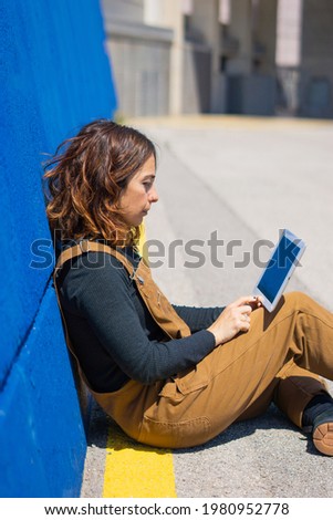 Girl working with tablet on the street sitting and leaning against a blue wall on the street on a sunny day.