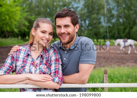 Couple on ranch. Happy young loving couple standing close to each other and smiling while standing on ranch with horse walking in the background