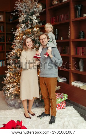 Winter holidays with family. Smiling Caucasian mother and father with baby girl daughter standing by decorated Christmas tree at home. Happy family celebrating Christmas or New Year together. 