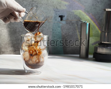 Hand in sterilized gloves pouring espresso over ice balls in double wall insulated glass on white marble background and rustic grey wall with rainbow light reflection. Refreshing summer drink theme.
