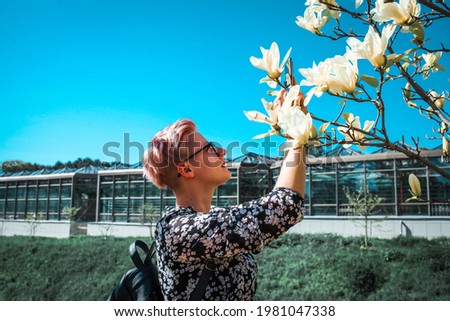 Positive blond woman traveler taking photo by camera under magnolia blossoms tree.