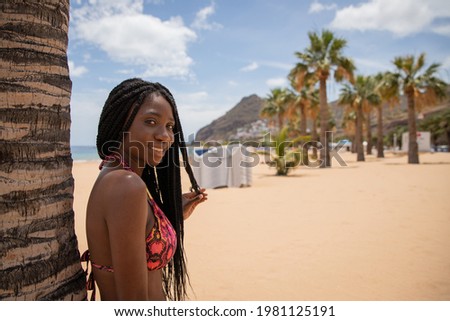 Attractive african girl enjoys her vacation on the beach, she leans her back on a palm tree and touches her braids with her hand.