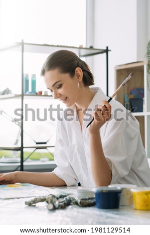 Home art. Painting hobby. Creative weekend. Profile portrait of inspired happy relaxed smiling lefthanded artist woman in white shirt with paintbrush in light studio.