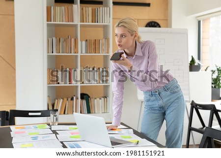 Confident businesswoman executive standing at work table with documents in office, recording audio message on phone, chatting with colleagues online or speakerphone, activating digital assistant
