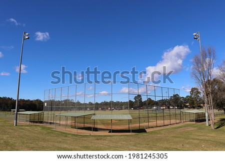 Suburban green grassed baseball field with metal fence, dug outs and tall flood light towers on a blue sky sunny day with small white clouds.