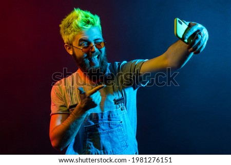 Handsome hipster bearded man wearing t shirt and denim overalls, broadcasting live stream, points to camera with finger, grimacing, winking and showing tongue. Colorful neon light, indoor studio shot