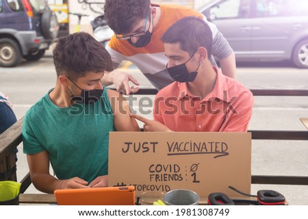 Three friends went for a drink to celebrate their coronavirus vaccination. They have a sign that reads just vaccinated, and the friends vs covid score. They point to the patch on the shoulder.