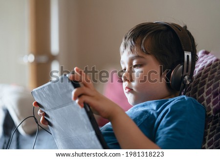 Child lying on couch wearing headphone listening to music, Close up face Kid sitting on sofa watching cartoons on tablet,Young boy playing game on touch pad or relaxing on his own in living room
