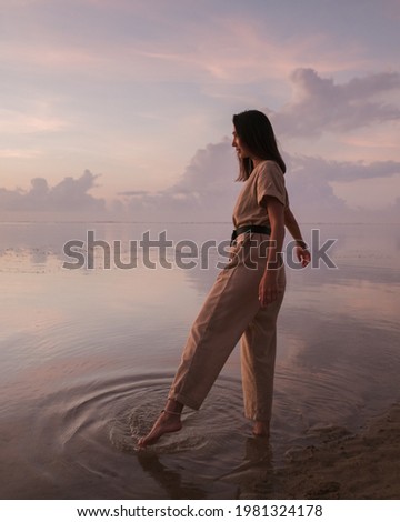 The girl touches the ocean with her foot and admires the beautiful sunrise on the beach.