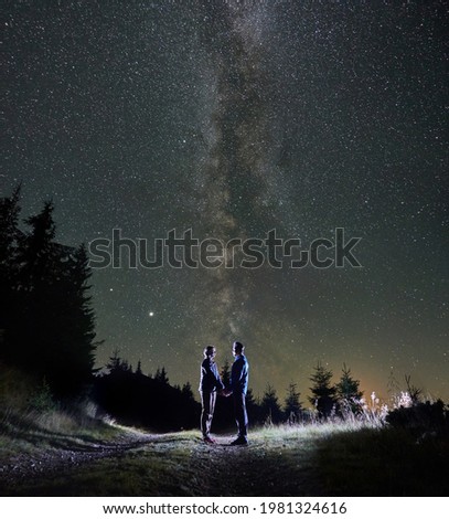 Magnificent view of night starry sky over mountain valley with couple travelers. Young woman and man holding hands while standing on mountain road under magical sky with stars and Milky Way.