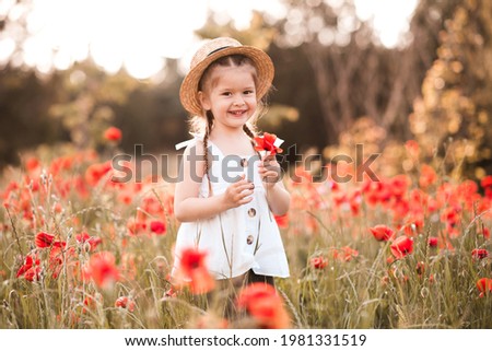 Smiling happy child girl 3-4 year old holding flower stand in poppy meadow outdoors over nature background. Wear white top and straw hat in field. Childhood. Summer season. Happiness. Freedom. Royalty-Free Stock Photo #1981331519