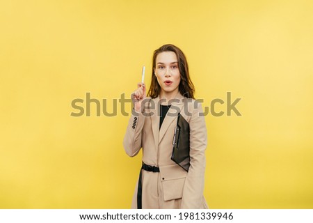 An attractive woman with a tablet and in a beige suit stands on a yellow background, looks at the camera and raises her hand with pen up, she has an idea.