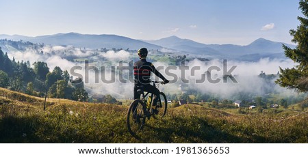 Man riding bicycle on grassy hill and looking at beautiful misty mountains. Male bicyclist enjoying panoramic view of majestic mountains during bicycle ride. Concept of sport, bicycling and nature. Royalty-Free Stock Photo #1981365653