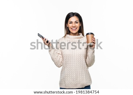Portrait of a smiling young woman holding mobile phone while drinking coffee and looking at camera isolated over white background