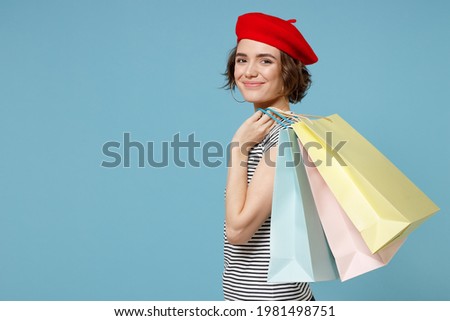 Side profile view of young woman 20s with short hairdo in french beret red hat striped t-shirt hold package bags with purchases after shopping isolated on pastel blue color background studio portrait
