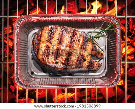 Roasted salmon steak in aluminum foil container on bbq grate over hot pieces of coals. Top view.