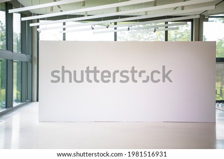 Modern gallery interior with blank white empty canvas. Contemporary, open space, city view and daylight. Marble floor. Green trees in background. Light spots,generic design furniture and building.