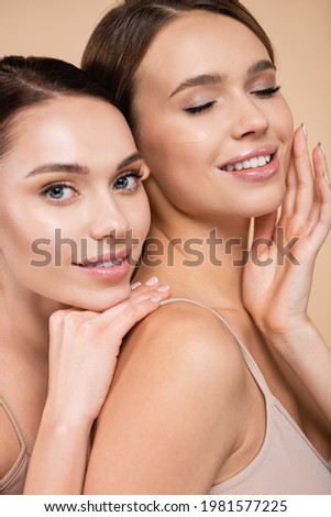 pleased woman touching face near friend looking at camera isolated on beige Royalty-Free Stock Photo #1981577225
