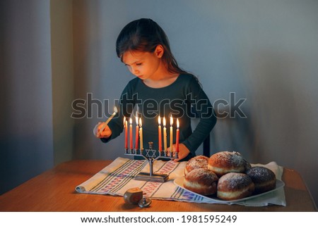 Traditional winter Jewish Hanukkah holiday. Girl lighting candles on menorah for at home. Child celebrating Chanukah festival of lights. Dreidel and Sufganiyot donuts on table. Israel Judaic holiday.