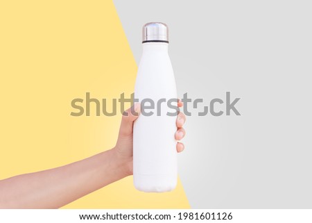 Close-up of female hand holding white reusable steel thermo water bottle isolated on two backgrounds of yellow and grey. Colors of the year 2021; ultimate gray and illuminating.