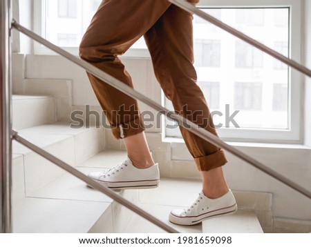 Woman in white sneakers and khaki trousers goes upstairs to her apartment. White staircase in apartment building. Casual outfit, urban fashion. Royalty-Free Stock Photo #1981659098