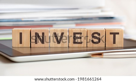 The word INVEST is written on wooden cubes standing on a tablet, in the background a stack of documents, selective focus. Can be used for business, education, financial, marketing concept.