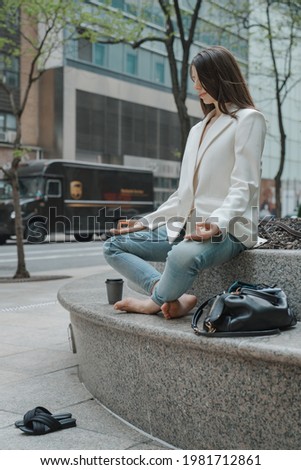Beautiful young adult woman meditating sitting crossed leg lotus pose in white jacket, blue jeans in New York city. Peaceful break time in busy city life 