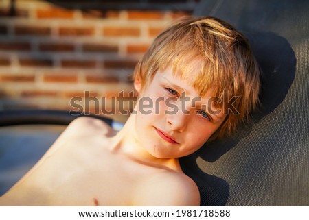 Blond 8 year old boy laying on a chair and sunbathing at home