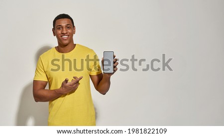 Cheerful young man in casual yellow t shirt smiling at camera, showing and pointing at smartphone with blank screen while standing isolated over gray background. Advertisement concept. Web Banner