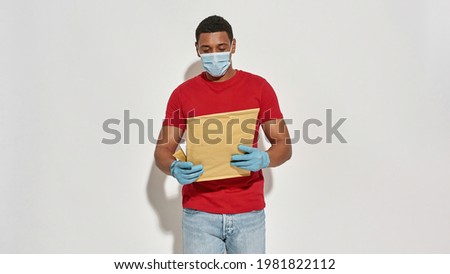 Delivery man wearing face mask and protective gloves holding, looking at parcels and envelops while posing isolated over light gray background. Service concept. Web Banner