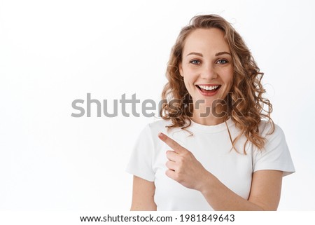 Cheerful blond girl with curly hairstyle, smiling broadly, pointing finger left at copy space, showing advertisement, white background