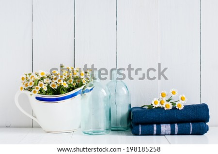 Chamomile flowers in vintage ceramic gravy boat, glass bottles and navy linen towels on white wooden background, home kitchen rustic decor