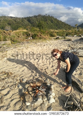 girl or young woman making camp fire on the beach, Sunset Sky at Gentle Annie, mussel is held in the front of the picture, New Zealand, west coast, south island