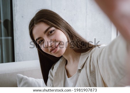 Blogger recording video or making online call. Portrait view of young millennial woman standing in living room, take self portrait for social networks on camera, smiling wide. Lifestyle concept