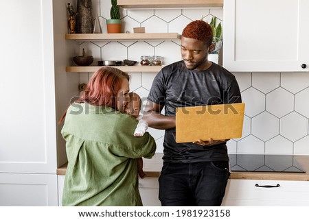 Young interracial family holding cute mixed-race baby Red haired young mother embracing lovingly standing in the modern kitchen, near Afro American father working online with yellow laptop in hands