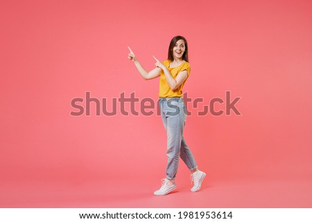 Full length portrait side view of amazed excited young brunette woman 20s in yellow casual t-shirt pointing index fingers aside up on mock up copy space isolated on pink color wall background studio Royalty-Free Stock Photo #1981953614