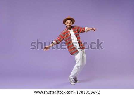Full length smiling young african american man in casual colorful shirt hat dancing standing on toes spreading hands isolated on violet background studio. People lifestyle concept. Mock up copy space. Royalty-Free Stock Photo #1981953692