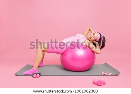 Positive active Asian woman leans over fitness ball has workout at home during quarantine poses on yoga mat dressed in sportswear isolated over pink background does pilates exercises. Sport training Royalty-Free Stock Photo #1981982300