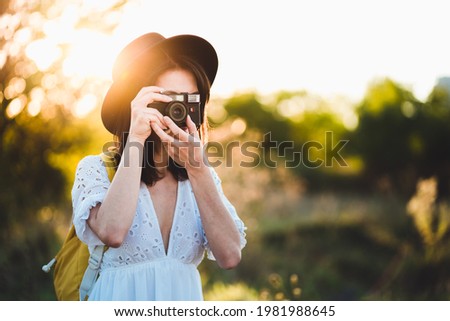 Hipster young woman tourist using vintage retro camera. Young fashionable female traveler with touristic backpack taking photos in nature at sunset. Majestic sunbeam from behind