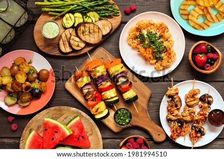 Healthy plant based summer bbq table scene. Top view on a dark wood background. Grilled fruit and vegetables, skewers, cauliflower steak and vegetarian sides. Meat substitute concept.