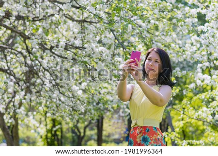 a young girl in a bright yellow-red summer dress takes pictures of herself on a smartphone against the background of a blooming apple orchard