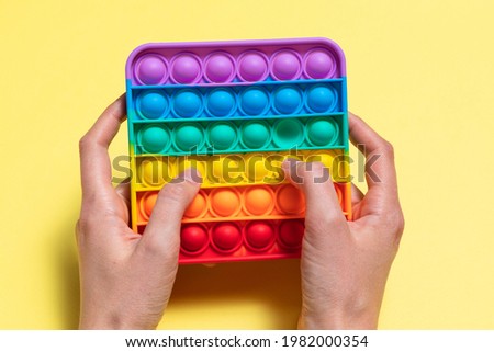 Kid's hands is holding a rainbow Pop It Toy against a yellow background. Trendy colorful fidget anti-stress sensory toy for popping bubbles. Push Pop Bubble Fidget Toy. Top view, copy space, close up
