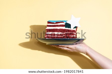 Female hand holding a plate with a piece of cake in the form of the USA flag on a yellow background, celebrating Independence Day, close up.