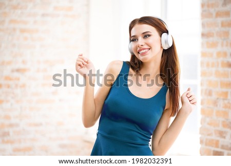 Happy smiling excited woman listening music in headphones at home. Beautiful girl in blue dress standing against window, indoors.