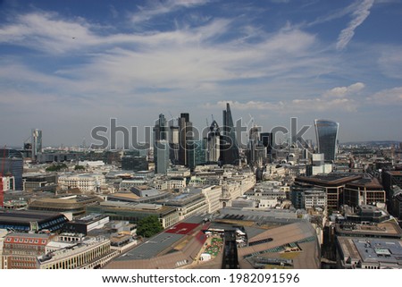Cityscape of London on a sunny day, roofs of various buildings fill the foreground, skyscrapers and other buildings form the skyline, blue sky with white cirrus and cumulus clouds is in the background