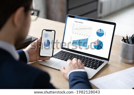 Business marketer using professional app for marketing data analyzing on laptop and smartphone screens, studying financial graphs, comparing diagrams on phone and computer. Analysis concept Royalty-Free Stock Photo #1982128043