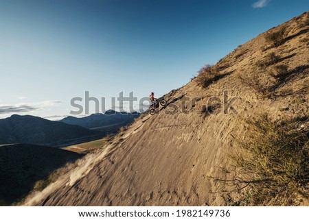 Extreme Rider climbing sand mountain top on off-road cross enduro motorcycle. Beautiful mountains landscape down on background, colourful autumn forest and river in sunshine 