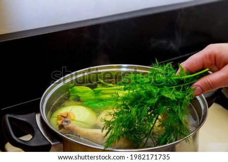 Woman put dill in the chicken broth with whole chicken and onion in steel pot.